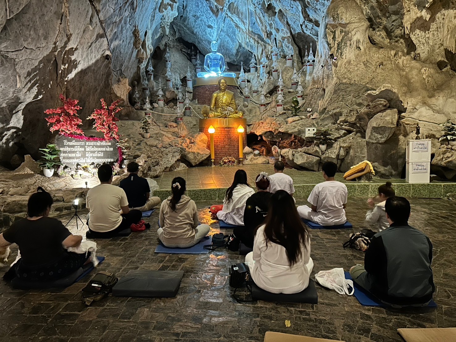 ผู้ปฏิบัติธรรมนั่งสมาธิหันหน้าเข้าหาพระประธานที่ประดับไฟภายในถ้ำ