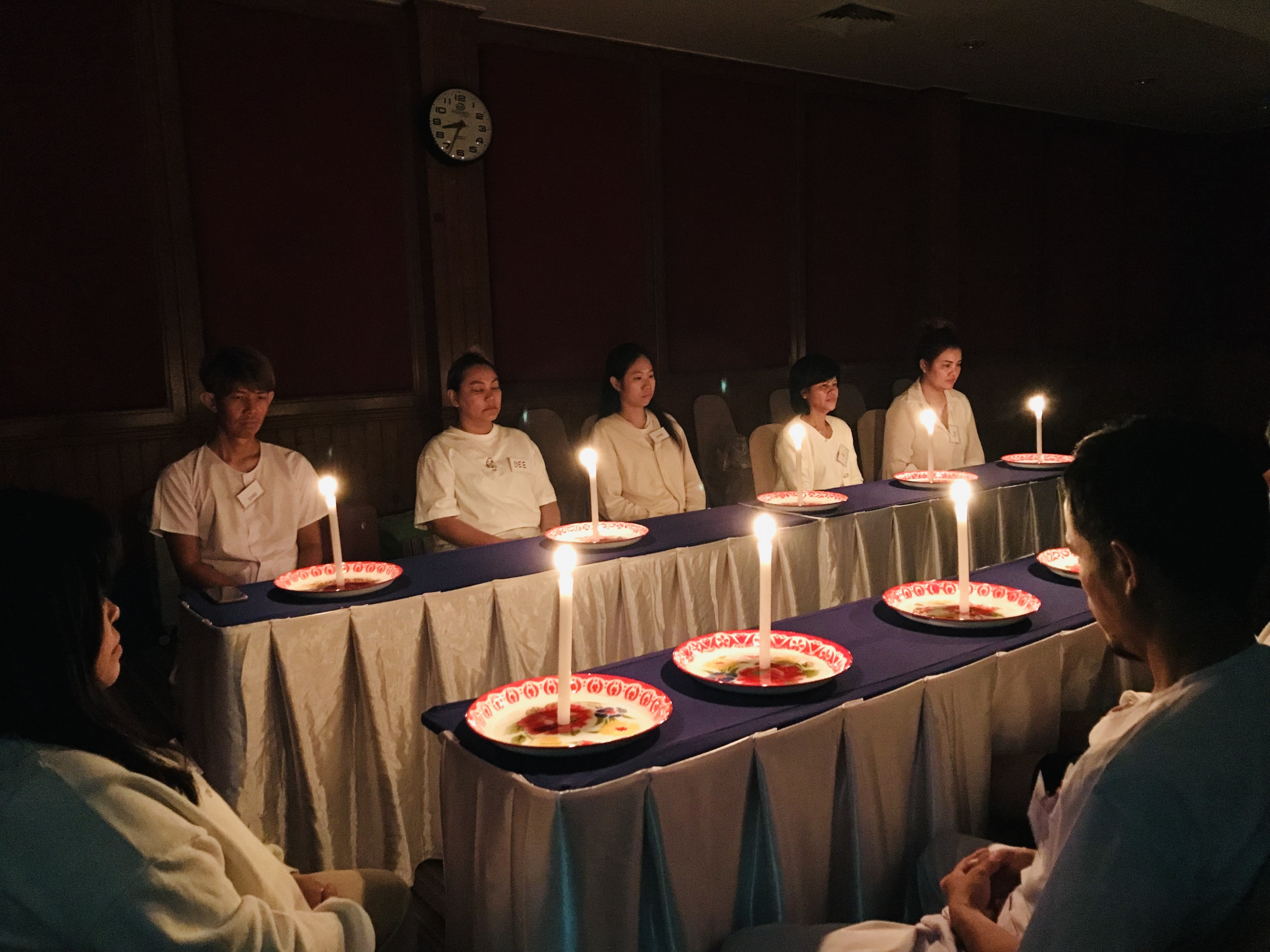 ผู้ปฏิบัติธรรมนั่งสำรวมรอบโต๊ะที่จุดเทียนวางบนจานในห้องแสงสลัว
